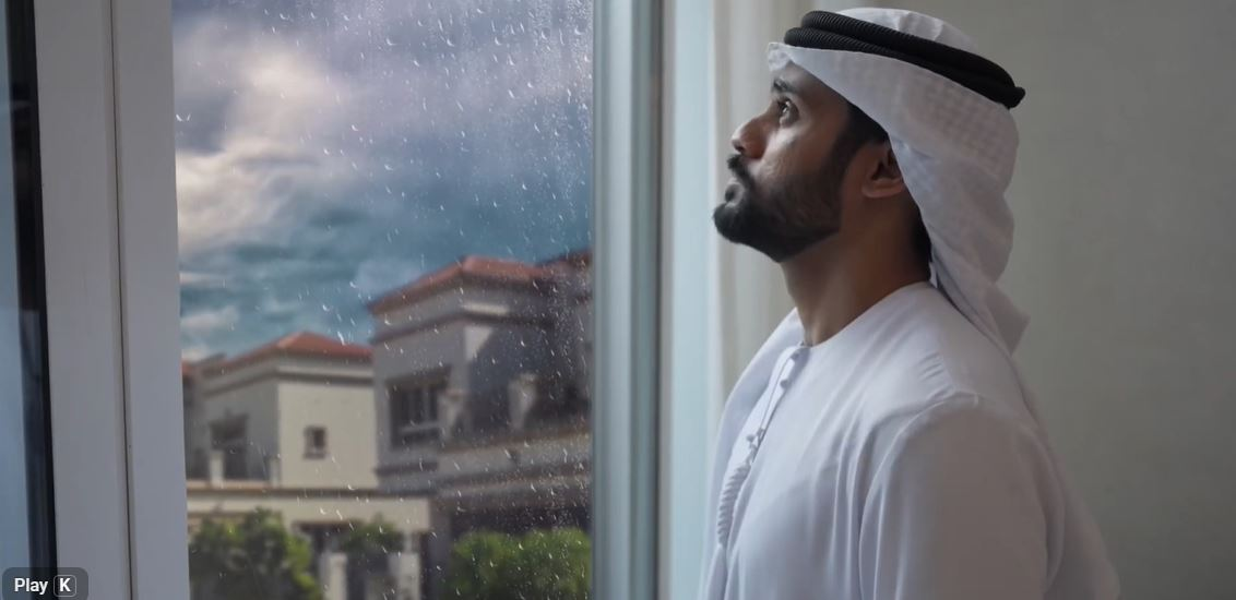 rainy-season-preparedness-dewa-uae.webp Man in traditional Emirati attire looking out a window on a rainy day during DEWA’s rainy season preparedness campaign.