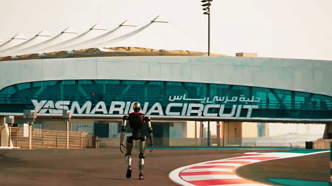 A humanoid autonomous robot walking on the track at Yas Marina Circuit with the iconic turquoise bridge structure in the background.